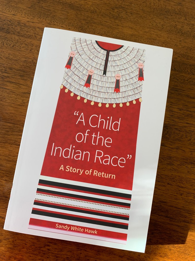 A book on a wooden table. The book is by Sandy White Hawk and the title is "A Child of the Indian Race: A Story of Return."